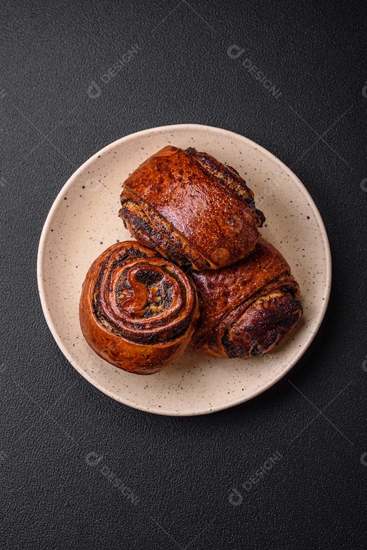 Deliciosos pães doces assados ​​com sementes de papoula e canela em uma placa de cerâmica sobre um fundo escuro de concreto