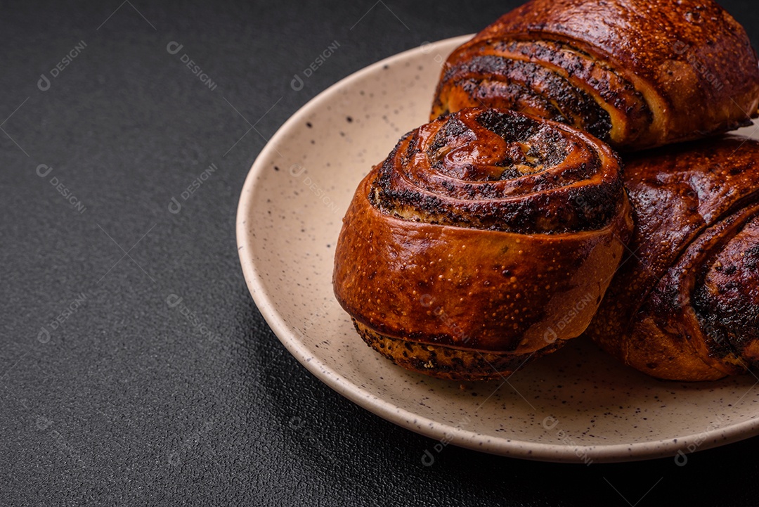 Deliciosos pães doces assados ​​com sementes de papoula e canela em uma placa de cerâmica sobre um fundo escuro de concreto