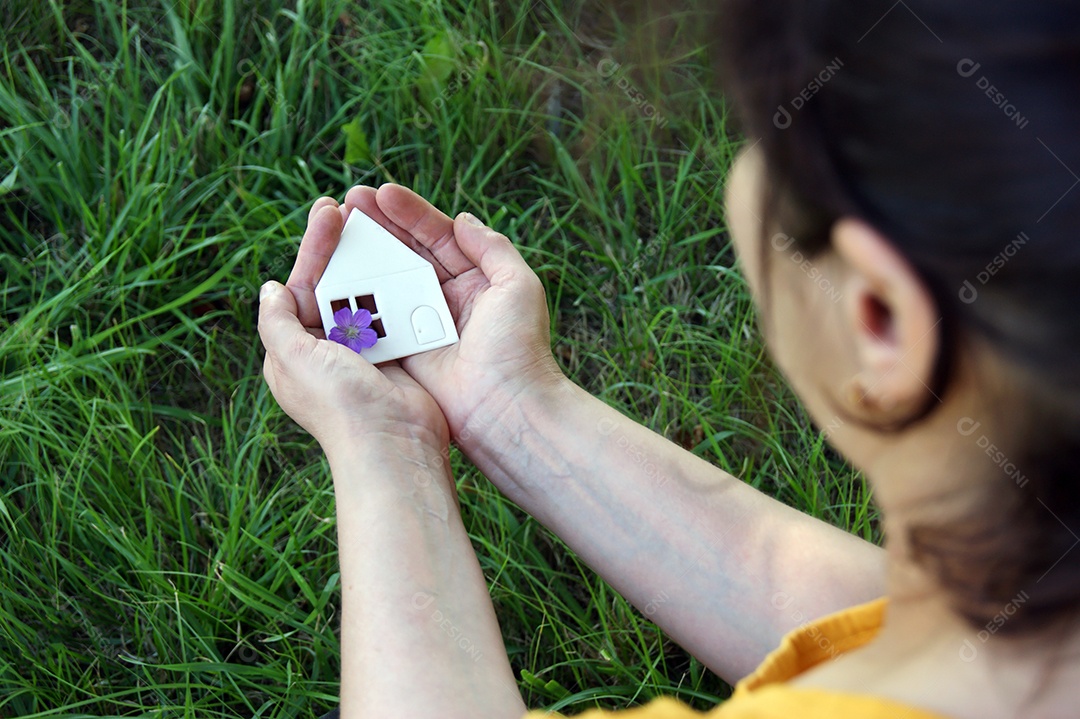Mulher segurando mini casa em mãos sobre fundo de um terreno verde