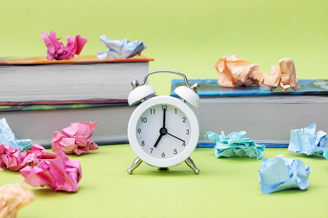 Despertador em frente a cadernos livros e papeis amassados