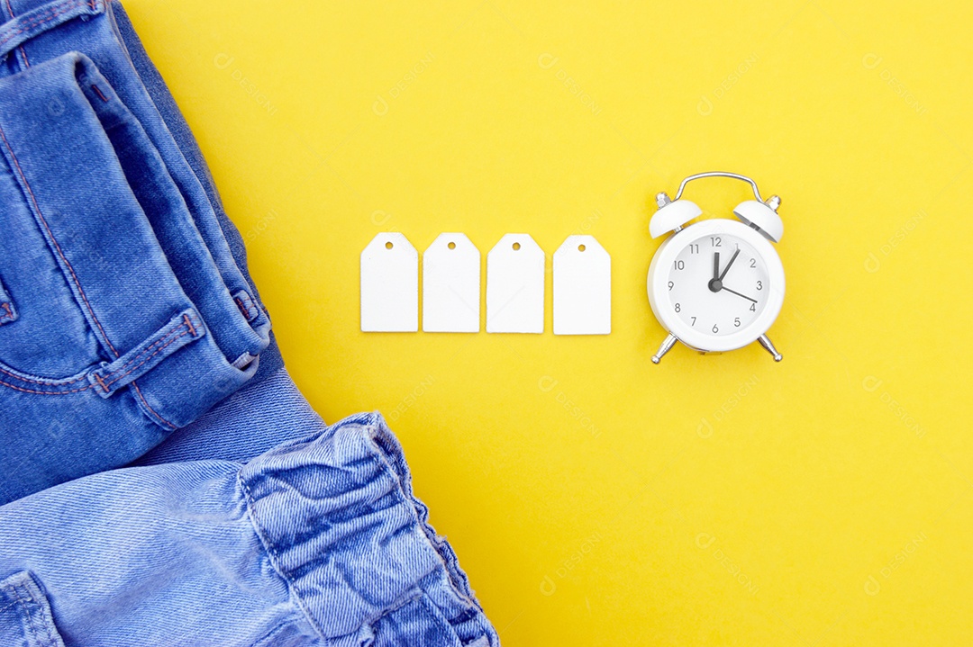 Roupas jeans azulado com etiquetas em branco e relógio sobre fundo amarelo