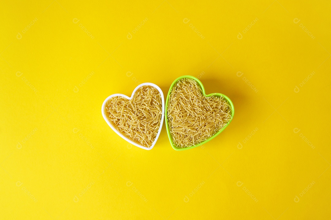 Duas tigelas de coração com macarrão cru amarelo sobre fundo amarelo claro