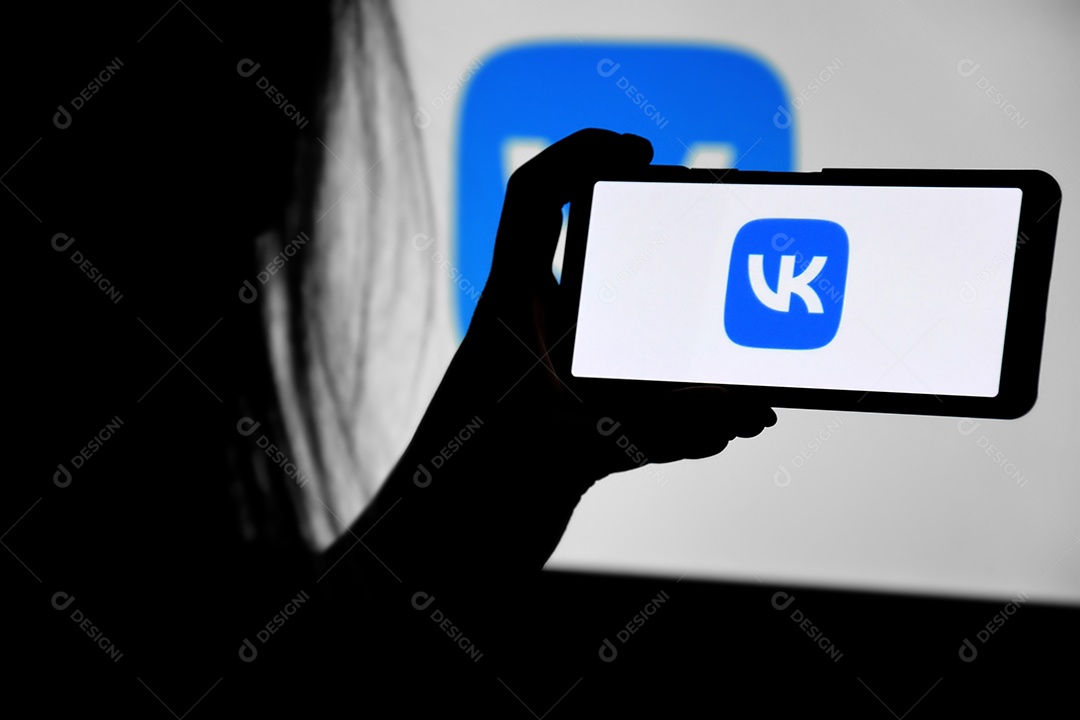 Um telefone com tela VK nas mãos de uma jovem contra o fundo de um monitor