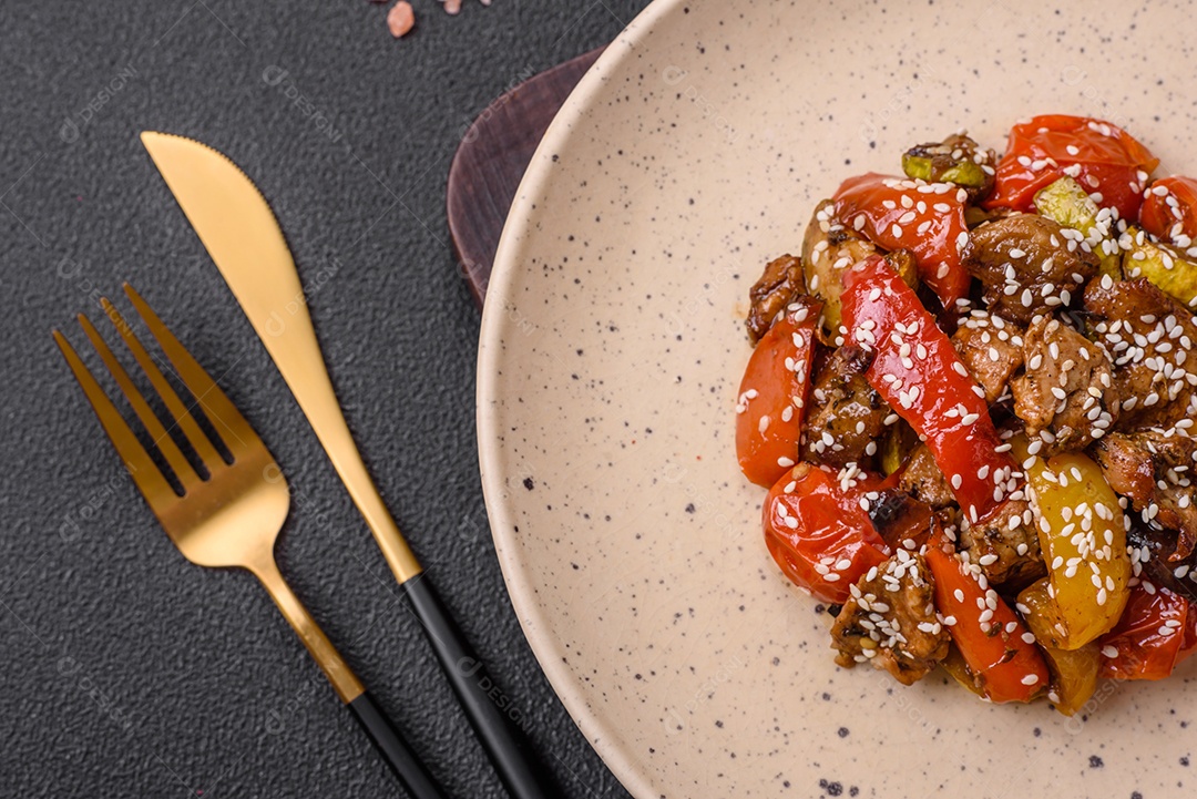 Salada quente com vitela, tomate, pimentão, abobrinha, gergelim, sal, especiarias e ervas sobre fundo escuro de concreto