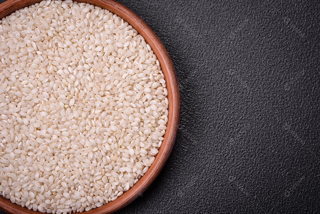 Arroz branco cru em uma tigela redonda de cerâmica sobre um fundo escuro de concreto. Cozinhar comida asiática