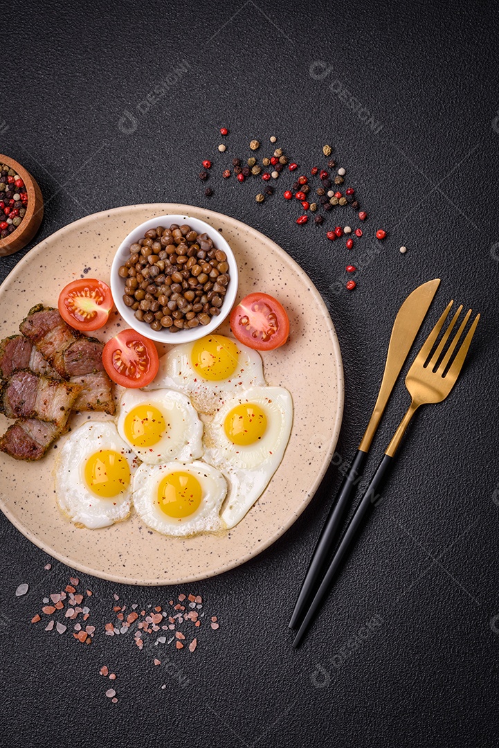 Delicioso café da manhã nutritivo com ovos de codorna fritos, bacon, legumes e tomate cereja com sal, especiarias e ervas em um fundo escuro de concreto
