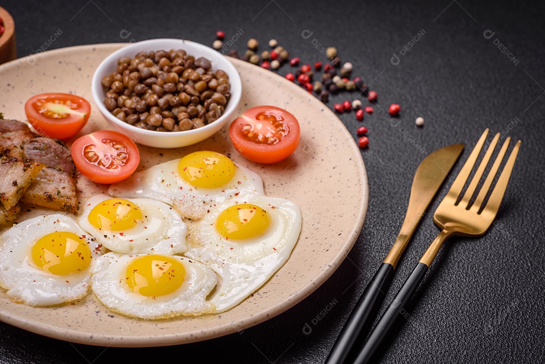 Delicioso café da manhã nutritivo com ovos de codorna fritos, bacon, legumes e tomate cereja com sal, especiarias e ervas em um fundo escuro de concreto