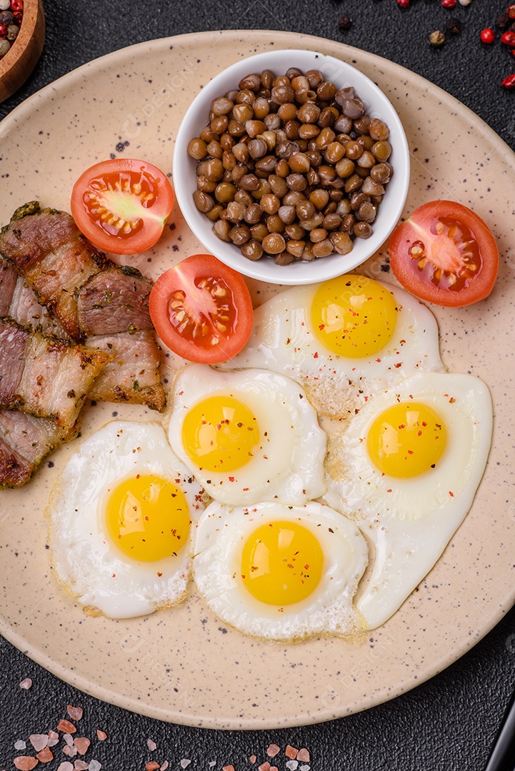 Delicioso café da manhã nutritivo com ovos de codorna fritos, bacon, legumes e tomate cereja com sal, especiarias e ervas em um fundo escuro de concreto