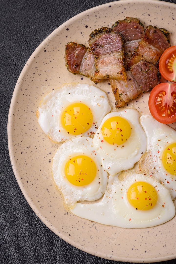 Delicioso café da manhã nutritivo com ovos de codorna fritos, bacon, legumes e tomate cereja com sal, especiarias e ervas em um fundo escuro de concreto