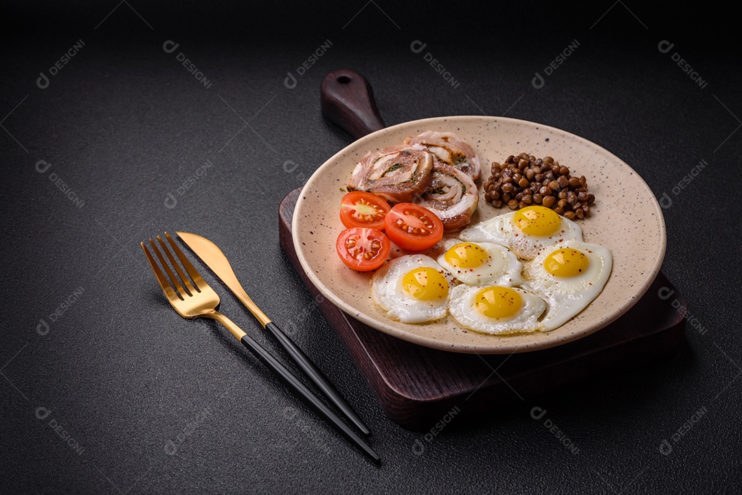 Delicioso café da manhã nutritivo com ovos de codorna fritos, bacon, legumes e tomate cereja com sal, especiarias e ervas em um fundo escuro de concreto