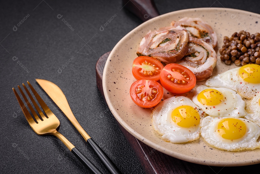 Delicioso café da manhã nutritivo com ovos de codorna fritos, bacon, legumes e tomate cereja com sal, especiarias e ervas em um fundo escuro de concreto