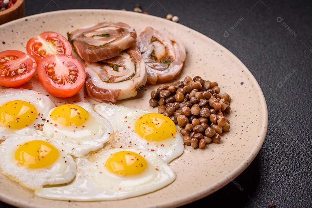 Delicioso café da manhã nutritivo com ovos de codorna fritos, bacon, legumes e tomate cereja com sal, especiarias e ervas em um fundo escuro de concreto