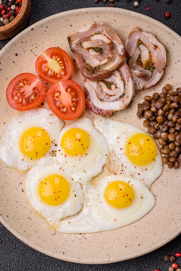 Delicioso café da manhã nutritivo com ovos de codorna fritos, bacon, legumes e tomate cereja com sal, especiarias e ervas em um fundo escuro de concreto