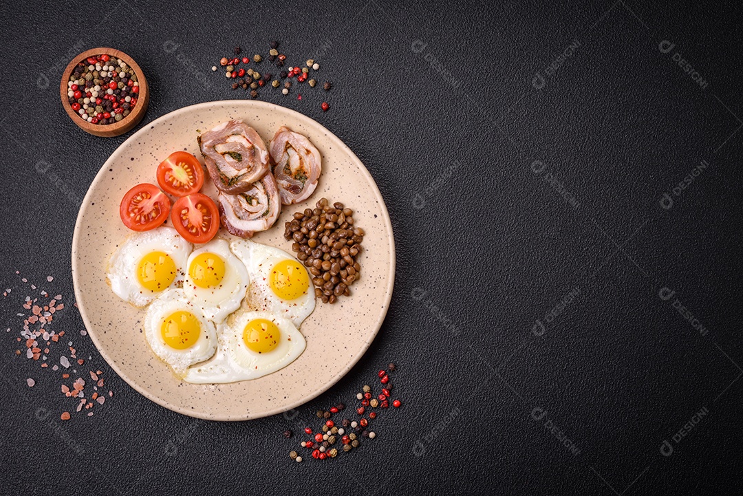 Delicioso café da manhã nutritivo com ovos de codorna fritos, bacon, legumes e tomate cereja com sal, especiarias e ervas em um fundo escuro de concreto