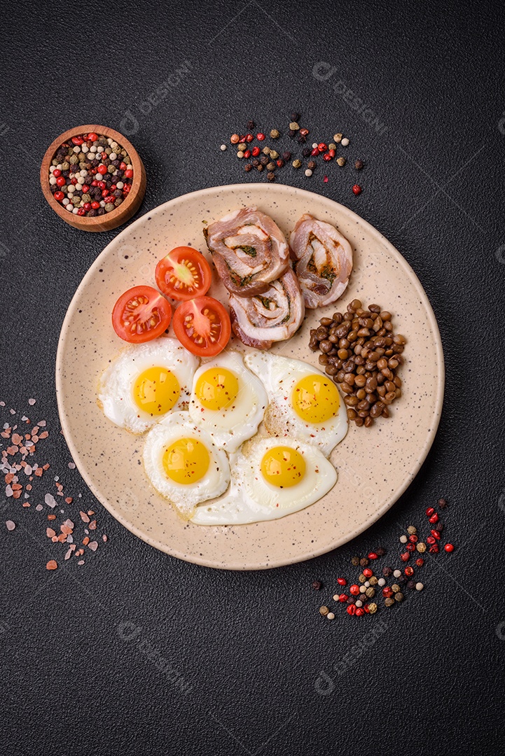 Delicioso café da manhã nutritivo com ovos de codorna fritos, bacon, legumes e tomate cereja com sal, especiarias e ervas em um fundo escuro de concreto