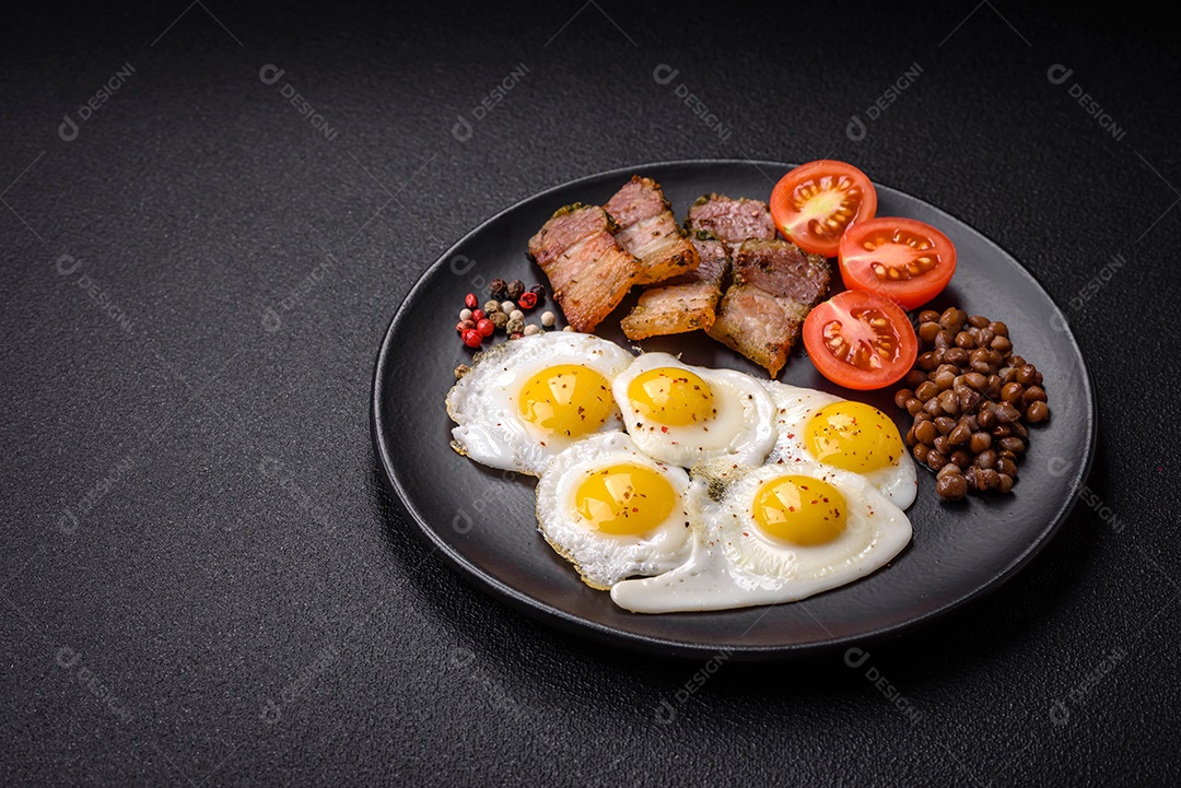 Delicioso café da manhã nutritivo com ovos de codorna fritos, bacon, legumes e tomate cereja com sal, especiarias e ervas em um fundo escuro de concreto