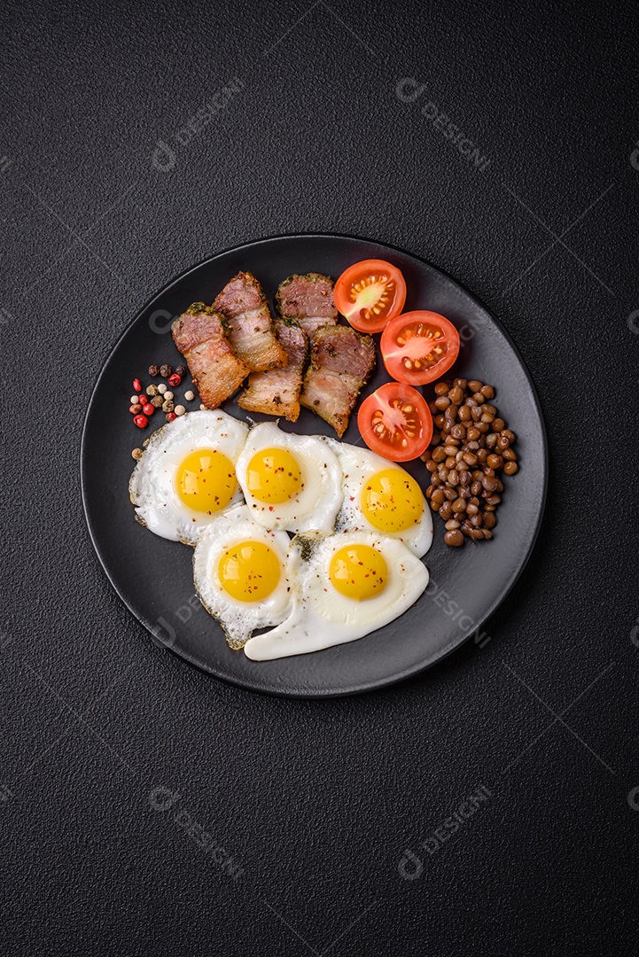 Delicioso café da manhã nutritivo com ovos de codorna fritos, bacon, legumes e tomate cereja com sal, especiarias e ervas em um fundo escuro de concreto