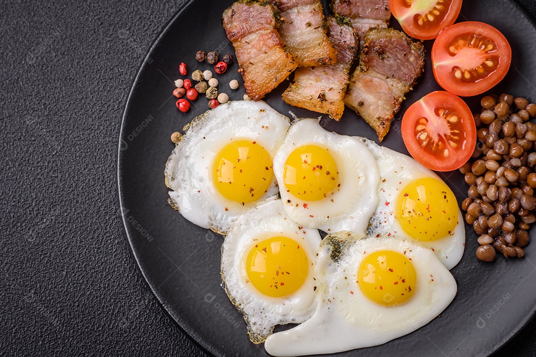 Delicioso café da manhã nutritivo com ovos de codorna fritos, bacon, legumes e tomate cereja com sal, especiarias e ervas em um fundo escuro de concreto