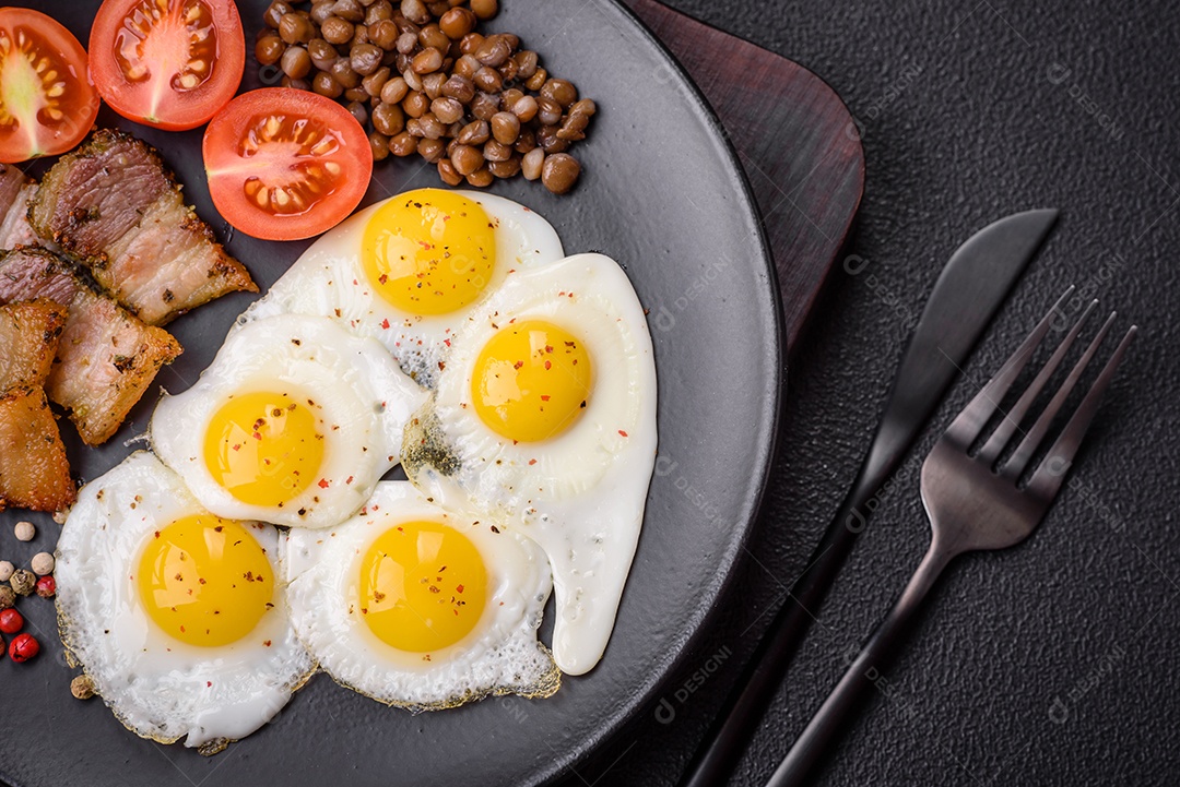 Delicioso café da manhã nutritivo com ovos de codorna fritos, bacon, legumes e tomate cereja com sal, especiarias e ervas em um fundo escuro de concreto