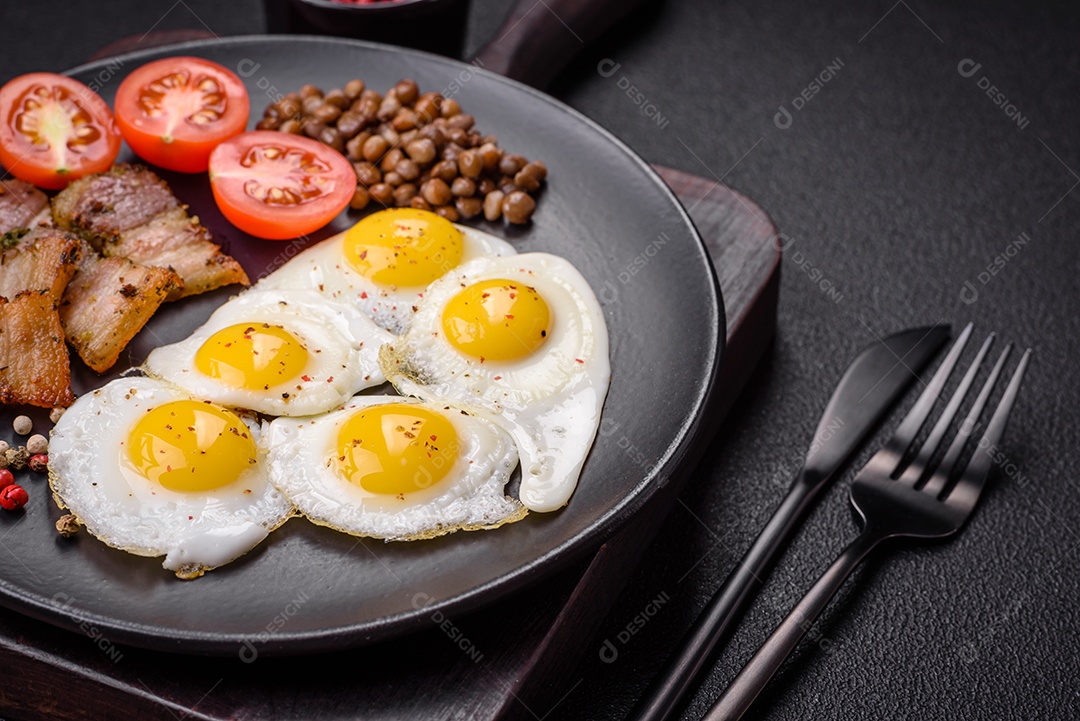 Delicioso café da manhã nutritivo com ovos de codorna fritos, bacon, legumes e tomate cereja com sal, especiarias e ervas em um fundo escuro de concreto