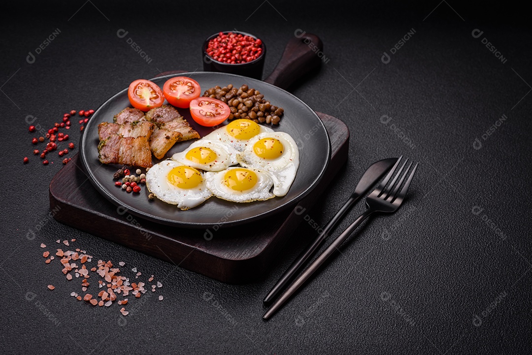 Delicioso café da manhã nutritivo com ovos de codorna fritos, bacon, legumes e tomate cereja com sal, especiarias e ervas em um fundo escuro de concreto