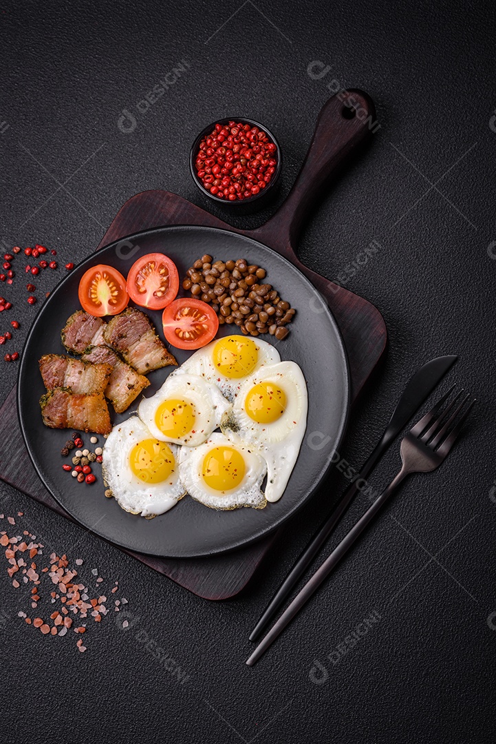 Delicioso café da manhã nutritivo com ovos de codorna fritos, bacon, legumes e tomate cereja com sal, especiarias e ervas em um fundo escuro de concreto