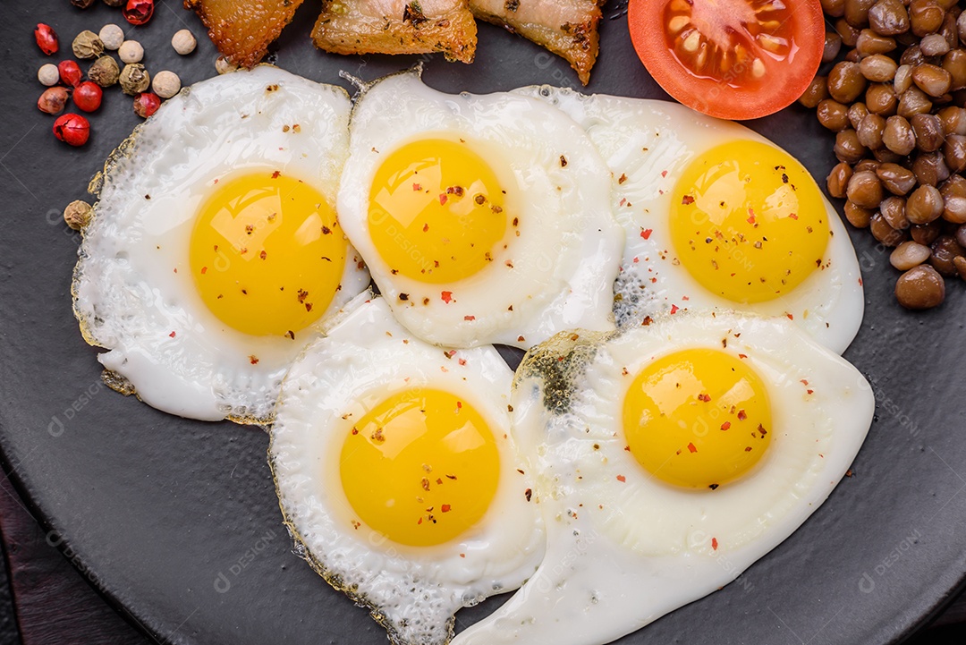 Delicioso café da manhã nutritivo com ovos de codorna fritos, bacon, legumes e tomate cereja com sal, especiarias e ervas em um fundo escuro de concreto