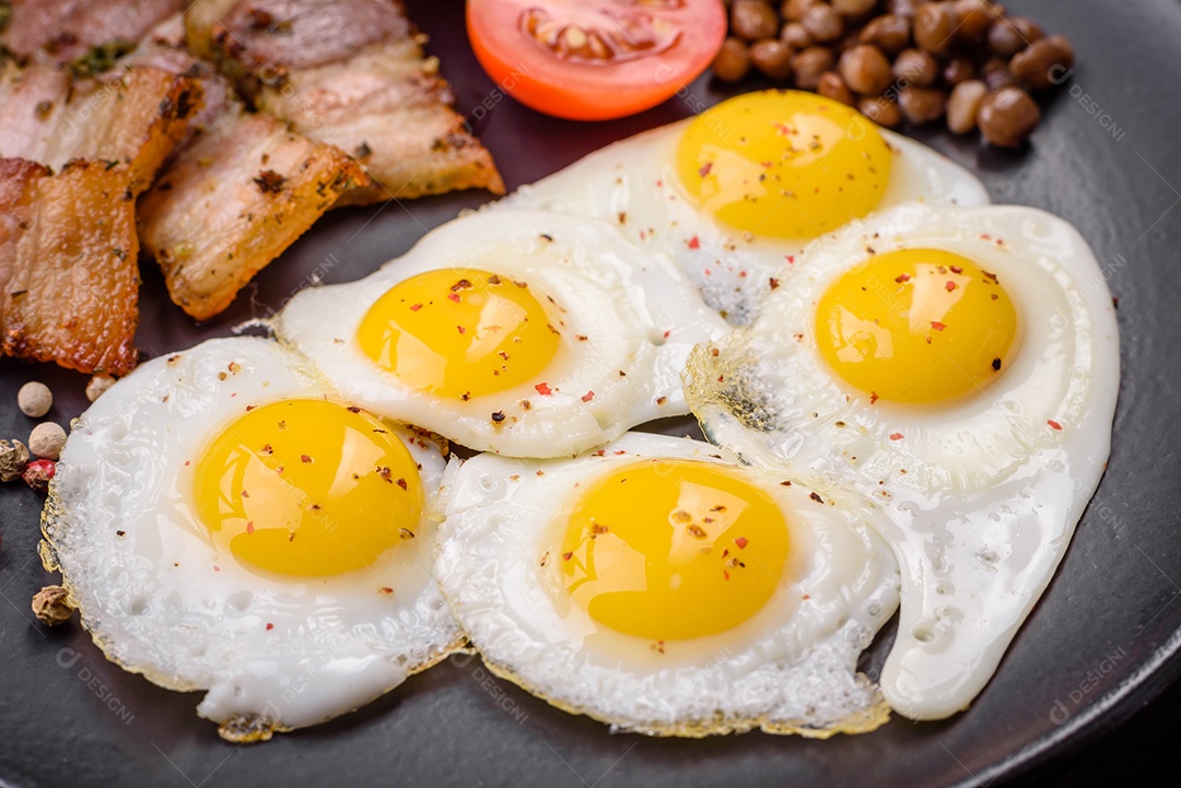 Delicioso café da manhã nutritivo com ovos de codorna fritos, bacon, legumes e tomate cereja com sal, especiarias e ervas em um fundo escuro de concreto