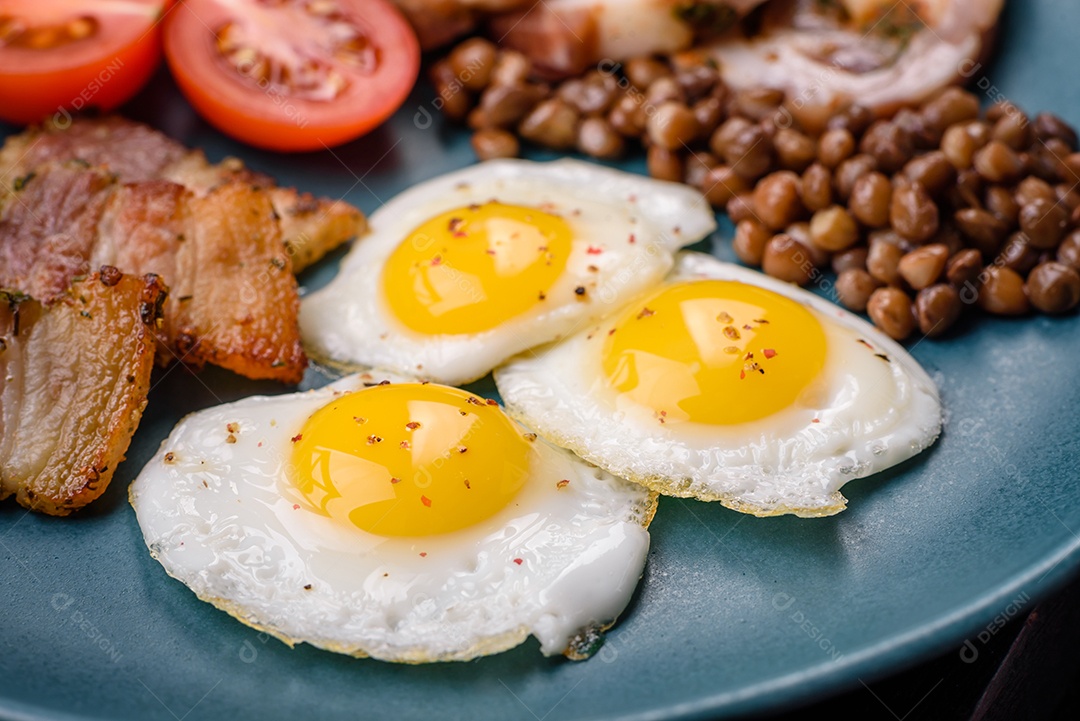 Delicioso café da manhã nutritivo com ovos de codorna fritos, bacon, legumes e tomate cereja com sal, especiarias e ervas em um fundo escuro de concreto