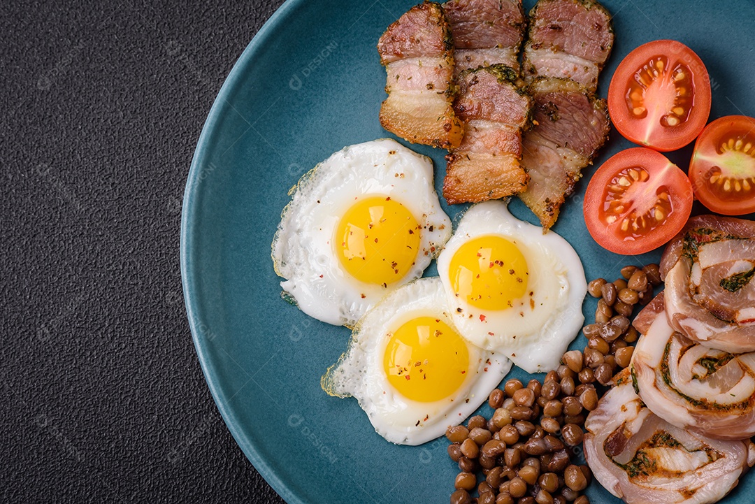 Delicioso café da manhã nutritivo com ovos de codorna fritos, bacon, legumes e tomate cereja com sal, especiarias e ervas em um fundo escuro de concreto