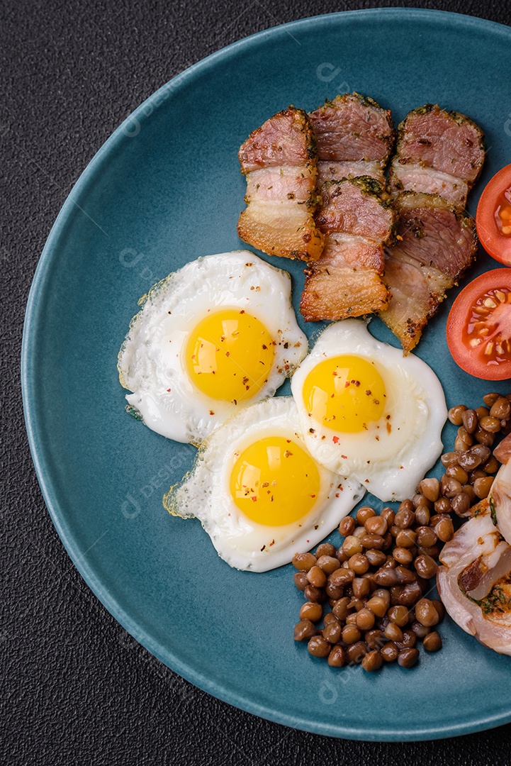 Delicioso café da manhã nutritivo com ovos de codorna fritos, bacon, legumes e tomate cereja com sal, especiarias e ervas em um fundo escuro de concreto