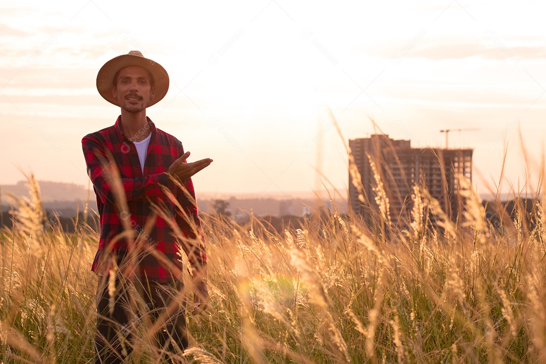 Agricultor com chapéu na plantação agrícola ao pôr do sol. Prédios e cidade desfocaram o fundo.