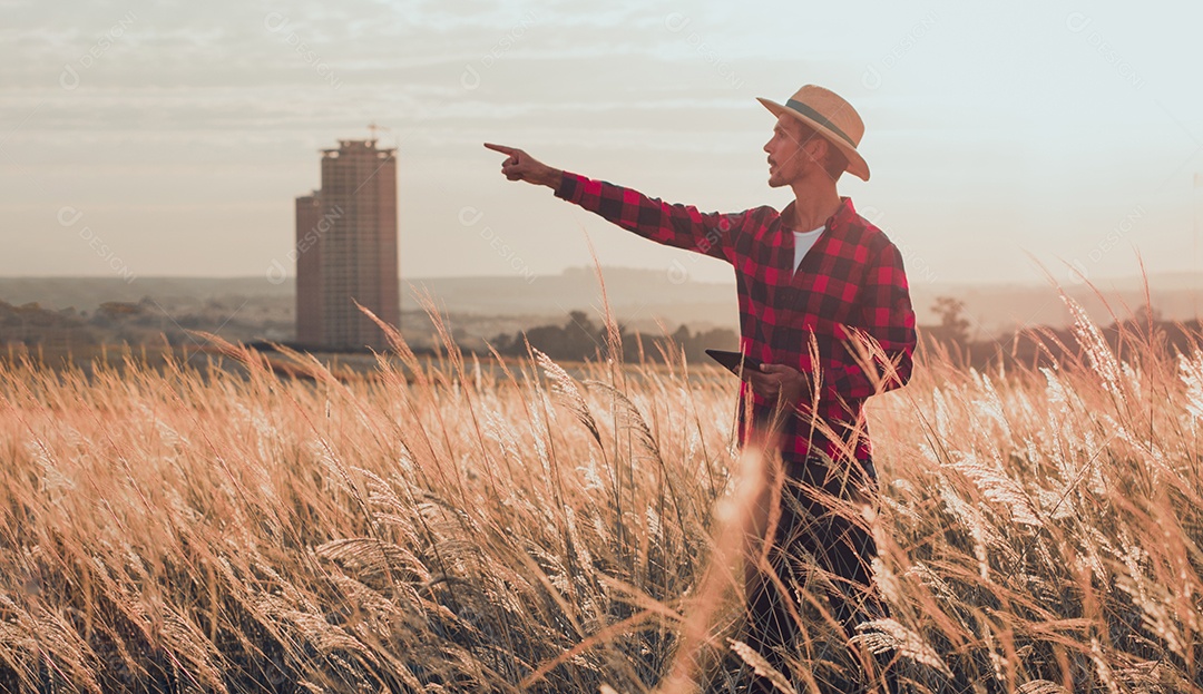 Agricultor com chapéu na plantação agrícola ao pôr do sol. Prédios e cidade desfocaram o fundo.