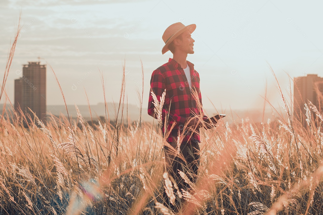 Agricultor com chapéu na plantação agrícola ao pôr do sol. Prédios e cidade desfocaram o fundo.
