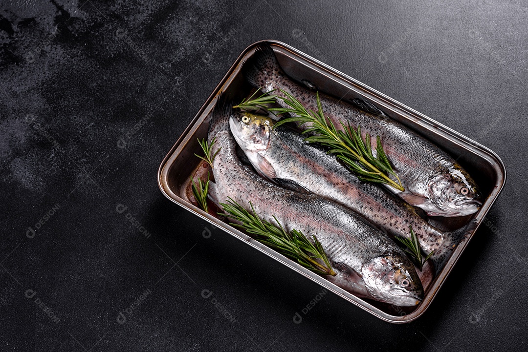 A truta crua está sobre uma tábua de pedra preta sobre uma mesa de concreto escuro. Perto estão: tomate, alecrim, limão, pimenta, cebola, sal