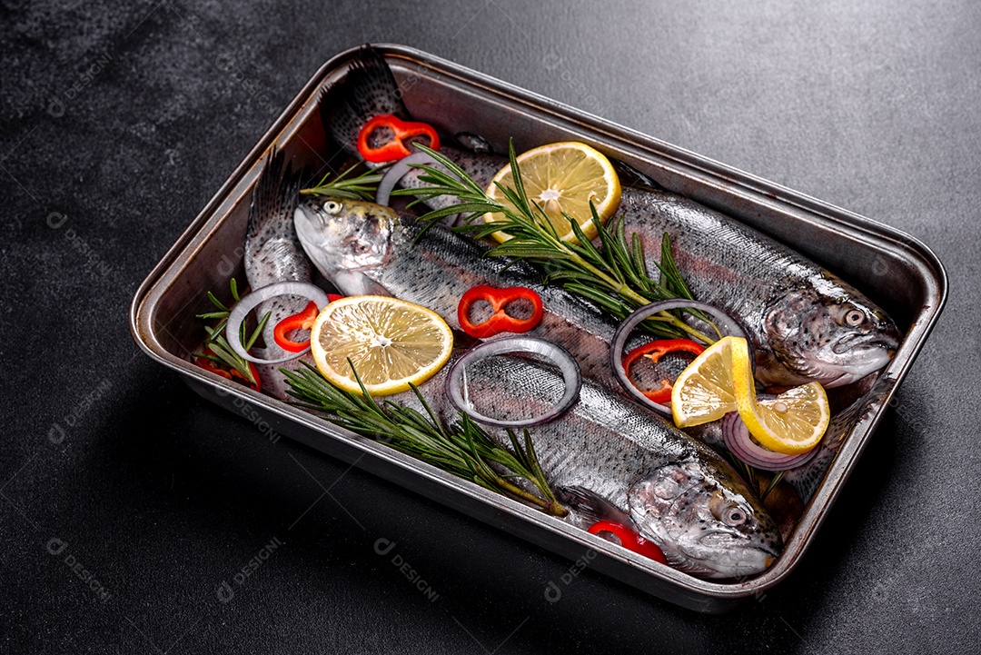 A truta crua está sobre uma tábua de pedra preta sobre uma mesa de concreto escuro. Perto estão: tomate, alecrim, limão, pimenta, cebola, sal