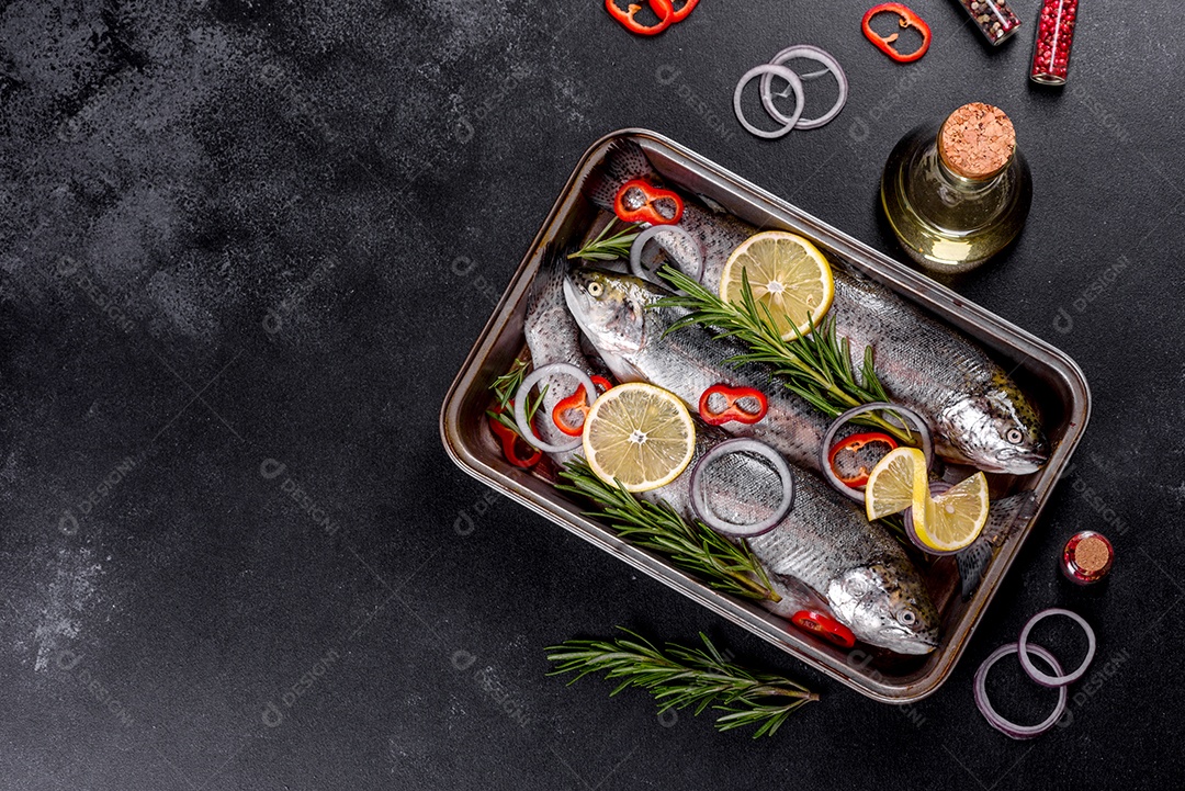 A truta crua está sobre uma tábua de pedra preta sobre uma mesa de concreto escuro. Perto estão: tomate, alecrim, limão, pimenta, cebola, sal