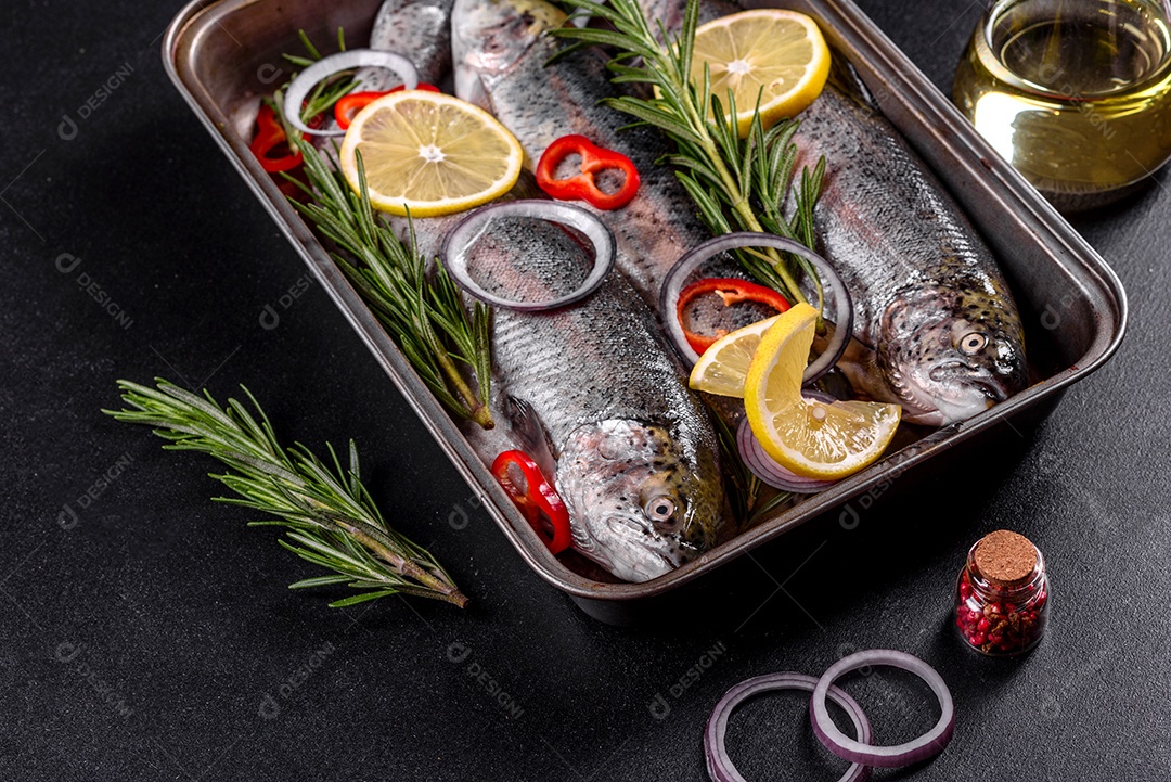 A truta crua está sobre uma tábua de pedra preta sobre uma mesa de concreto escuro. Perto estão: tomate, alecrim, limão, pimenta, cebola, sal
