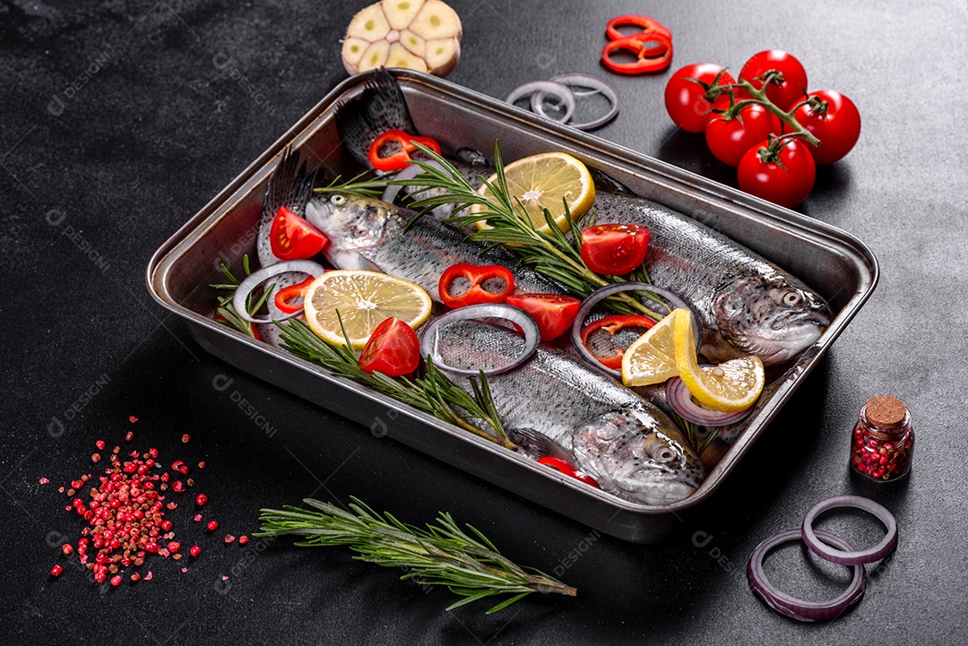 A truta crua está sobre uma tábua de pedra preta sobre uma mesa de concreto escuro. Perto estão: tomate, alecrim, limão, pimenta, cebola, sal