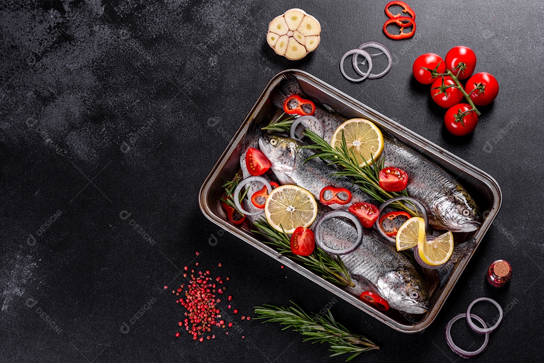 A truta crua está sobre uma tábua de pedra preta sobre uma mesa de concreto escuro. Perto estão: tomate, alecrim, limão, pimenta, cebola, sal