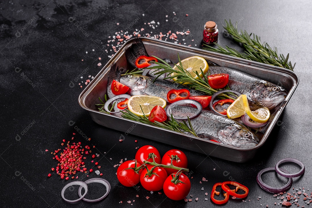 A truta crua está sobre uma tábua de pedra preta sobre uma mesa de concreto escuro. Perto estão: tomate, alecrim, limão, pimenta, cebola, sal