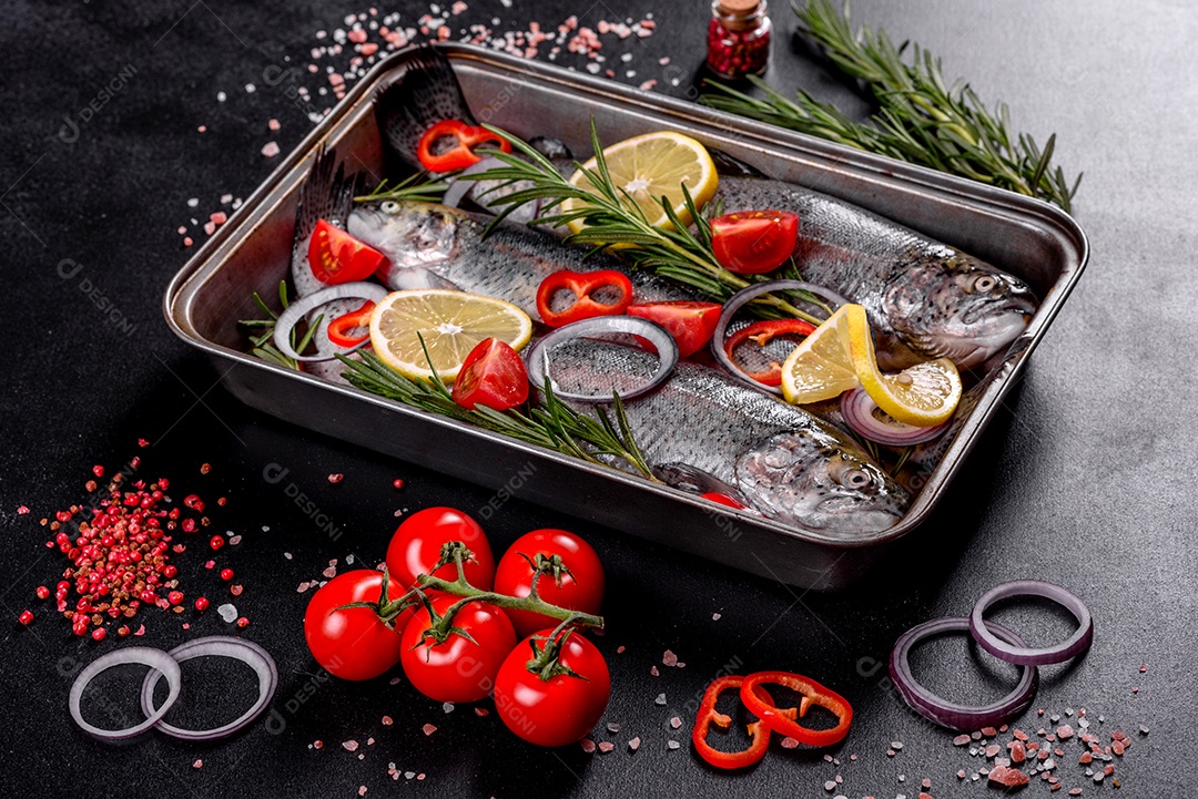 A truta crua está sobre uma tábua de pedra preta sobre uma mesa de concreto escuro. Perto estão: tomate, alecrim, limão, pimenta, cebola, sal