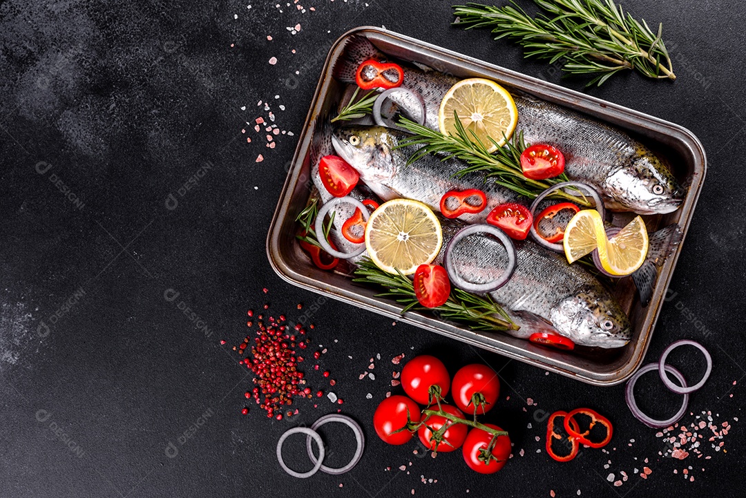 A truta crua está sobre uma tábua de pedra preta sobre uma mesa de concreto escuro. Perto estão: tomate, alecrim, limão, pimenta, cebola, sal