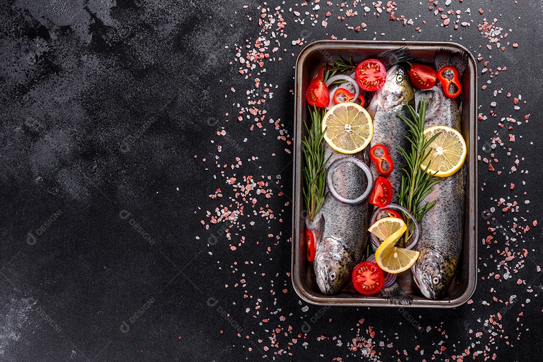 A truta crua está sobre uma tábua de pedra preta sobre uma mesa de concreto escuro. Perto estão: tomate, alecrim, limão, pimenta, cebola, sal