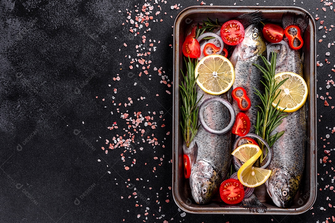 A truta crua está sobre uma tábua de pedra preta sobre uma mesa de concreto escuro. Perto estão: tomate, alecrim, limão, pimenta, cebola, sal