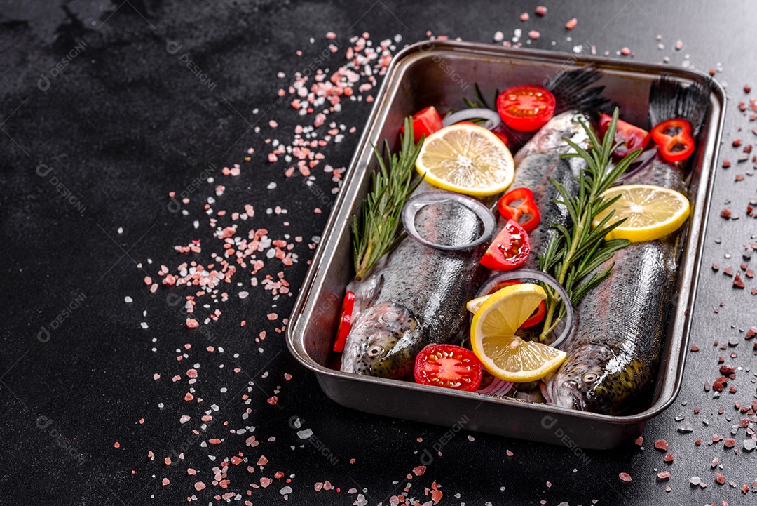 A truta crua está sobre uma tábua de pedra preta sobre uma mesa de concreto escuro. Perto estão: tomate, alecrim, limão, pimenta, cebola, sal