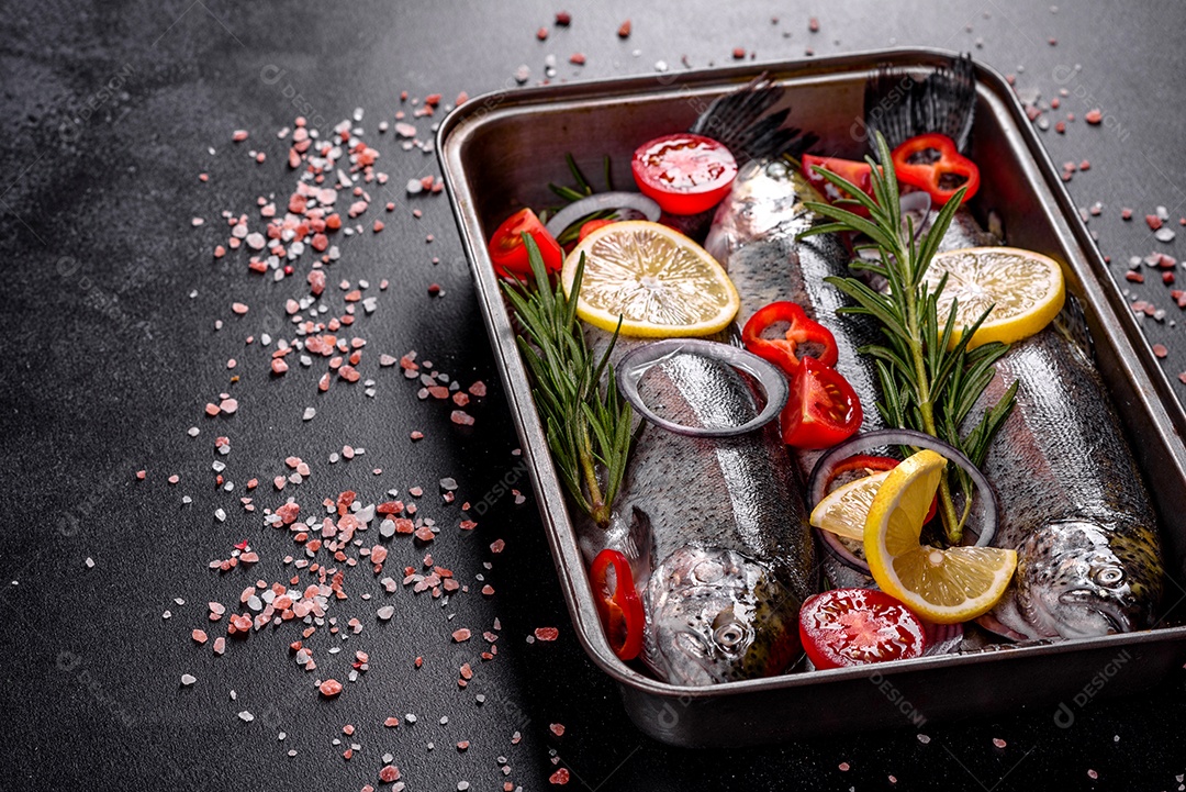 A truta crua está sobre uma tábua de pedra preta sobre uma mesa de concreto escuro. Perto estão: tomate, alecrim, limão, pimenta, cebola, sal