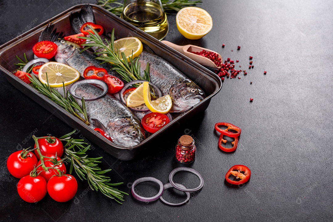 A truta crua está sobre uma tábua de pedra preta sobre uma mesa de concreto escuro. Perto estão: tomate, alecrim, limão, pimenta, cebola, sal