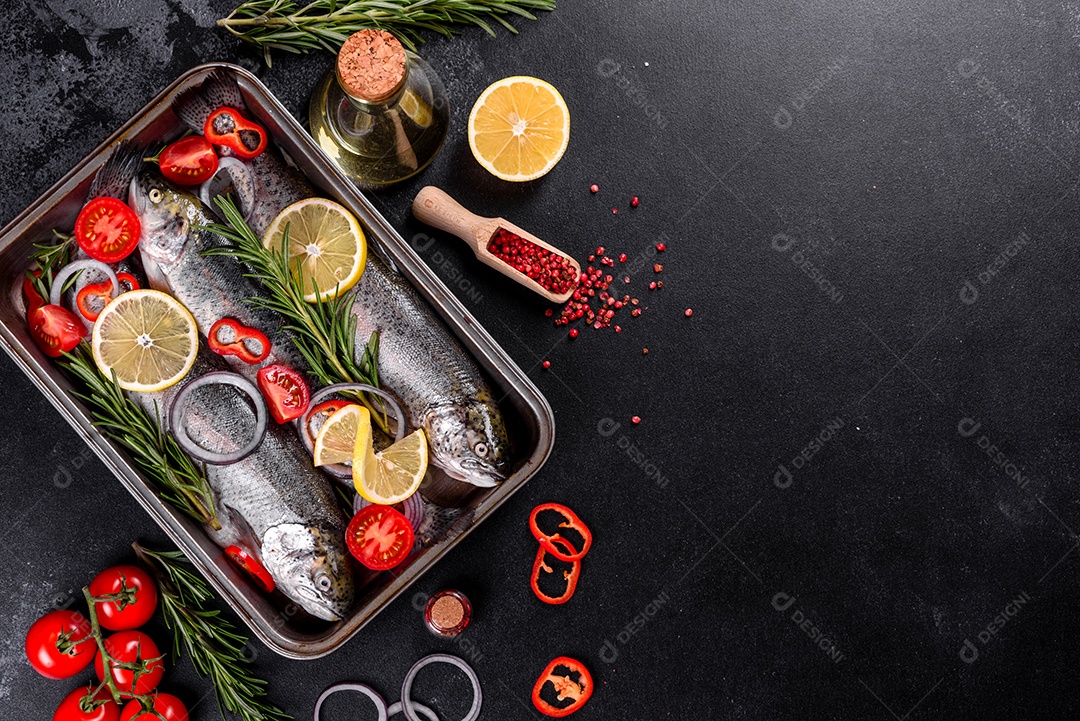 A truta crua está sobre uma tábua de pedra preta sobre uma mesa de concreto escuro. Perto estão: tomate, alecrim, limão, pimenta, cebola, sal