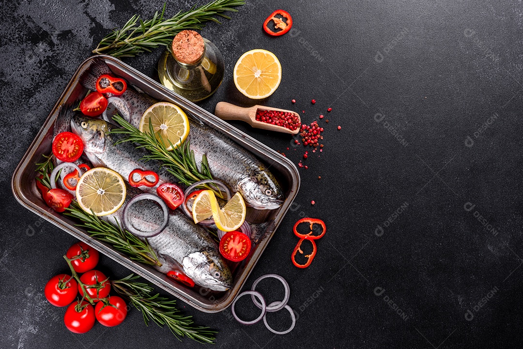 A truta crua está sobre uma tábua de pedra preta sobre uma mesa de concreto escuro. Perto estão: tomate, alecrim, limão, pimenta, cebola, sal